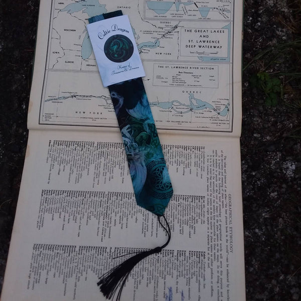 A decorative bookmark with a black tassel and a blue-green patterned body rests on an open vintage book. The bookmark has a white paper sleeve attached near the top with a circular logo and the text 'Celtic Dragon, Keeper of Secrets & Dreams'.