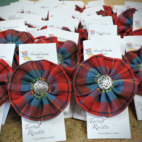 Several handmade British Columbia tartan rosettes are attached to white cards labeled 'Tartan Rosette' and arranged on a tabletop. The rosettes have a pleated fabric look with pewter Celtic knot center medallion.