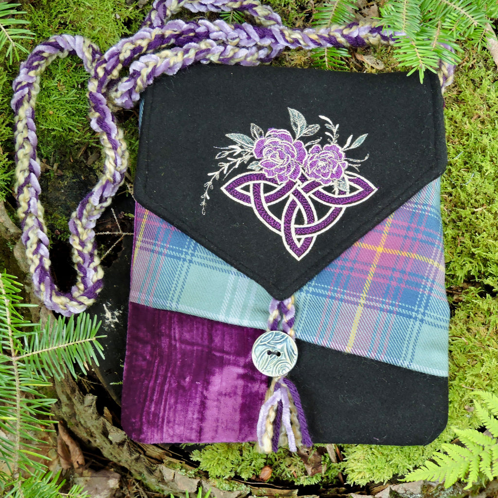 A handmade shoulder bag made from patchwork fabrics, including black wool, purple velvet, Hame tartan, and a purple braided strap, displayed on a bed of green pine needles and moss.