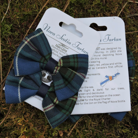 A handmade Nova Scotia Tartan hair bow in a classic green, red, and white plaid pattern, featuring a central silver-toned metal lion rampant emblem. The bow is attached to a white cardboard display card with historical text about the Nova Scotia flag, and is photographed lying on a natural mossy ground background.