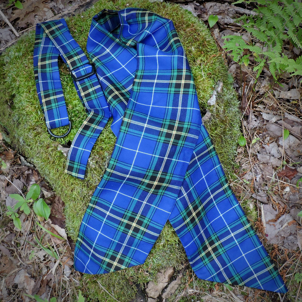 A NS tartan wreath sash and matching hanger laid out on a mossy log outdoors, surrounded by leaves and green plants.