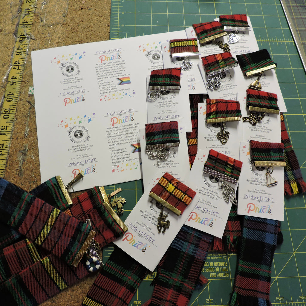 Pride Tartan keychains displayed on a sheet of paper with text on a green cutting mat.