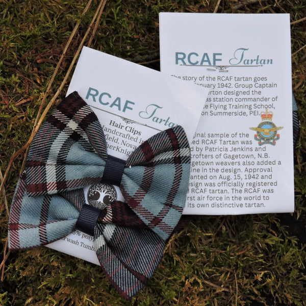 A handmade RCAF Tartan hair bow in a light blue, navy, and red plaid pattern, styled on top of printed historical cards and a green grass background.
