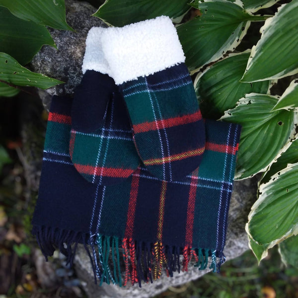 A pair of RCMP tartant mittens  and scarf with a white faux-fur cuff, displayed on a rock surrounded by green hosta leaves. 