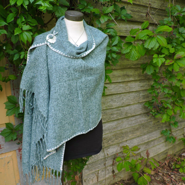 Mannequin wearing a green poncho with fringe against a wooden wall and greenery.